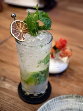 A refreshing Mojito cocktail or mocktail served in tall highball glass. The drink is packed with crushed ice, muddled fresh green mint leaves, and topped with a generous sprig of mint and an artistic dried lime wheel garnish. The fizzy, clear beverag