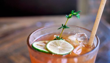 Kafe ortamında ahşap masada servis edilen ferahlatıcı bir bardak buzlu limon çayı ya da şeftali çayı. Kehribar renkli içecek taze yeşil limon dilimleri ve narin bir kekik filizi ile süslenerek aromatik bitkisel bir dokunuş eklenir. Çevre dostu saman kağıdı