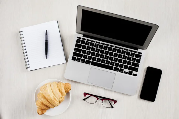 Laptop, smartphone, croissant, glasses and notebook — Stock Photo, Image