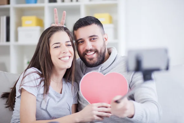 Joyeux jeune beau couple prenant des photos avec téléphone intelligent sur selfie stick. Jeune couple s'amusant dans le salon — Photo