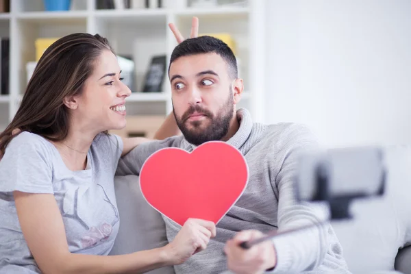 Feliz joven hermosa pareja tomando fotos con teléfono inteligente en palo selfie. Pareja joven divirtiéndose en el salón —  Fotos de Stock