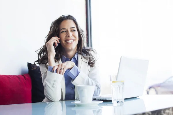 Wanita bisnis muda yang duduk di sebelah jendela mengerjakan laptop dan berbicara di telepon — Stok Foto