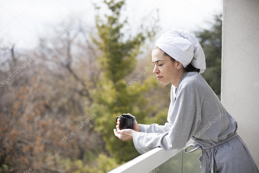 Young woman in bathrobe drinking coffee on the balcony in the morning