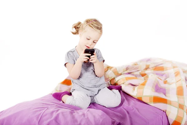 Cute little blonde girl under the pillow laughing and lying on the bed with purple linen — Stock Photo, Image