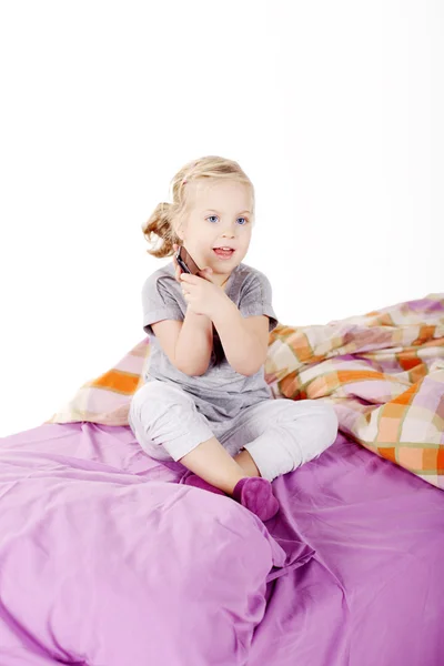 Linda niña rubia debajo de la almohada riendo y acostada en la cama con lino morado — Foto de Stock