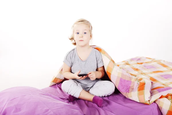 Cute little blonde girl under the pillow laughing and lying on the bed with purple linen — Stock Photo, Image