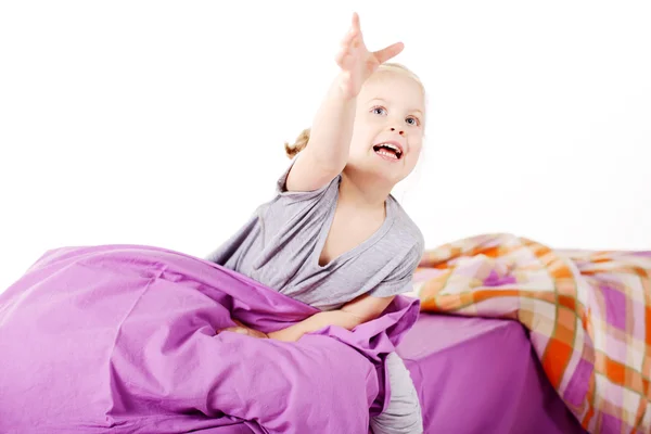 Cute little blonde girl under the pillow laughing and lying on the bed with purple linen Royalty Free Stock Images