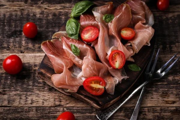 Thin slices of prosciutto with mixed with basil, cherry tomatoes on wooden cutting board,vintage background — Stock Photo, Image