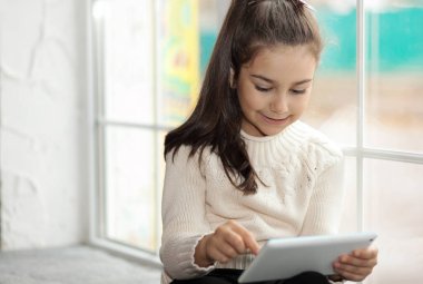 Evdeki pencerenin önünde otururken tablet kullanan tatlı küçük kız.