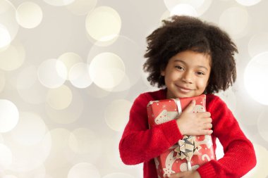 Gülümseyen, kıvırcık saçlı küçük bir kızın portresi. Elinde bir Noel hediyesi var. 
