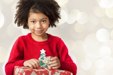 Gülümseyen, kıvırcık saçlı küçük bir kızın portresi. Elinde bir Noel hediyesi var. 