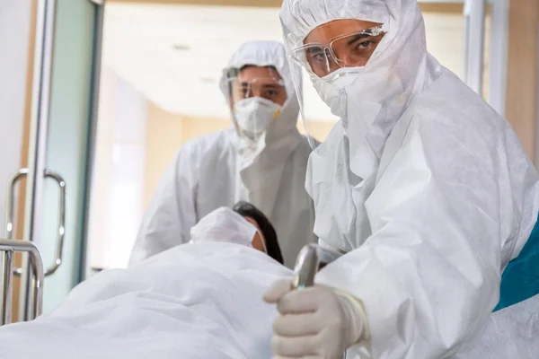 Asyalı hemşire ve doktor takım elbise ve yüz maskesi takarak sedye yatağı ve ciddi şekilde enfekte olmuş koronavirüs ya da kovid 19 hastası hastanedeki karantina odasına itiyorlar..