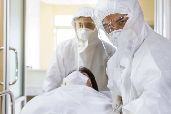 Asyalı hemşire ve doktor takım elbise ve yüz maskesi takarak sedye yatağı ve ciddi şekilde enfekte olmuş koronavirüs ya da kovid 19 hastası hastanedeki karantina odasına itiyorlar..