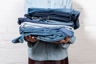 cropped view of woman holding stack of blue denim clothes near white brick wall clipart