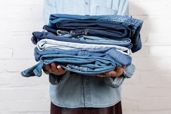 cropped view of woman holding stack of blue denim clothes near white brick wall