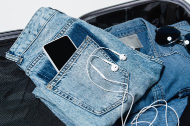 Kulaklıkların yanındaki kot pantolonların cebindeki akıllı telefonun üst görüntüsü, kot gömlek ve güneş gözlükleri seyahat çantasında paketlenmiş.