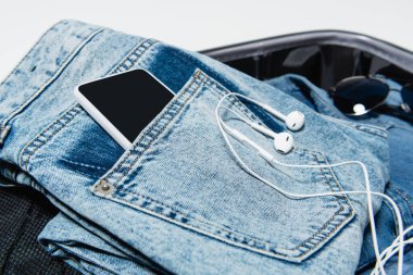 close up view of smartphone in pocket of jeans near earphones and sunglasses in travel bag clipart