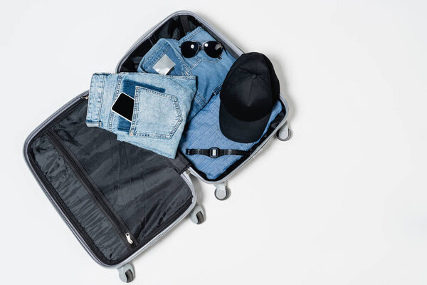 top view of suitcase packed with denim garments, black cap, smartphone, sunglasses and electronic wristwatch on white