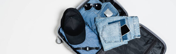 top view of mobile phone in pocket of jeans near denim shirt, black cap, electronic wristwatch and sunglasses in suitcase on white, banner