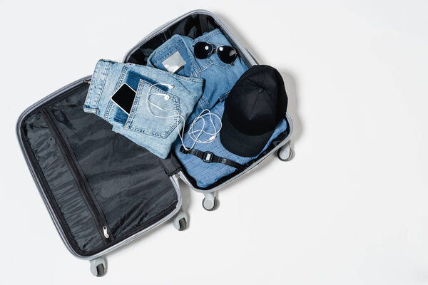 top view of suitcase with denim clothes, mobile phone, black cap, electronic wristwatch, sunglasses and earphones on white