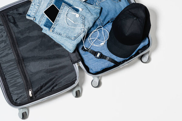 top view of travel bag with blue denim garments, smartphone, earphones, black cap and electronic wristwatch on white