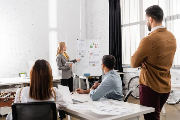 Dijital tabletli iş kadını, bulanık önplanda çok ırklı meslektaşlarının yakınındaki grafiklere işaret ediyor 