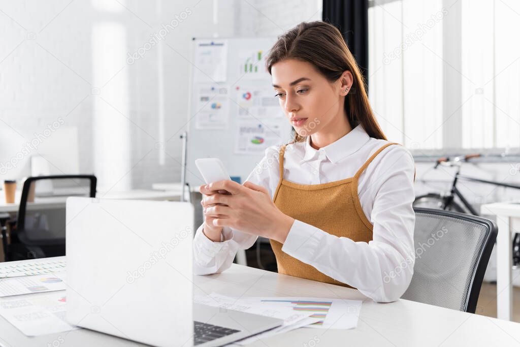Young businesswoman using smartphone near documents and laptop on blurred foreground 