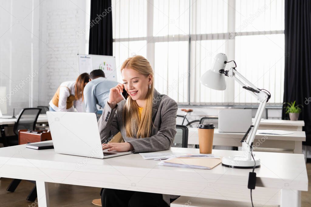 Smiling businesswoman talking on smartphone near laptop and coffee to go in office 