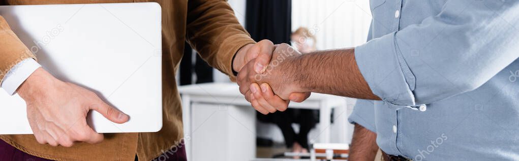 Cropped view of businessmen with laptop handshaking in office, banner 