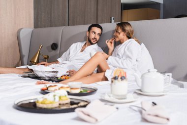 Genç ırklar arası bornozlu çift otel yatağında bulanık önplanda lezzetli yiyeceklerin yanında oturuyor. 