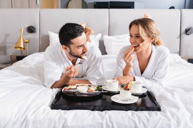 Genç ırklar arası bornozlu çift otel yatağında kahvaltı yapıyorlar. 