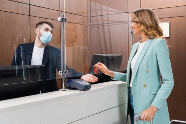Oteldeki tıbbi maskeli resepsiyonistin yanında elinde anahtar tutan neşeli kadın. 