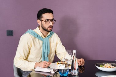 Muslim man in eyeglasses sitting near food and water on table in hotel room  clipart
