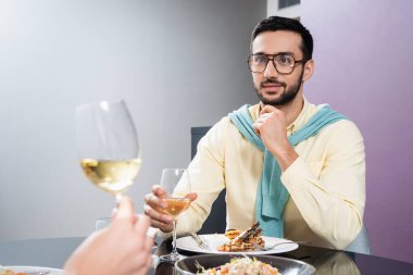 Kız arkadaşının yanında şarap bardağı tutan Arap bir adam ve otelde bulanık bir ön planda akşam yemeği. 