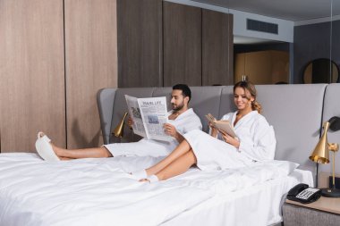 Otelde elinde gazeteyle Arap erkek arkadaşının yanında kitap okuyan gülümseyen bir kadın. 