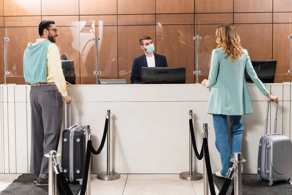 ホテルロビーの医療マスクのマネージャーの近くに立ってスーツケースを持つ異人種間のカップル — ストック写真