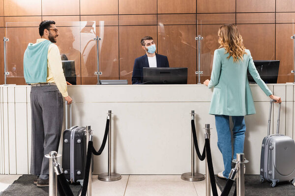 Interracial couple with suitcases standing near manager in medical mask in hotel lobby 