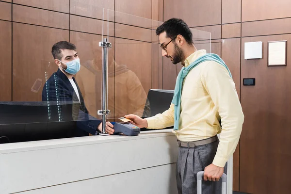 Receptionist in medical mask holding payment terminal near muslim man with suitcase and credit card in hotel lobby