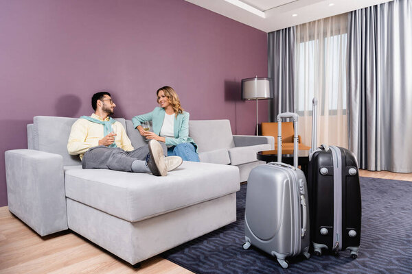 Smiling woman with wine looking at muslim boyfriend near suitcases in hotel 
