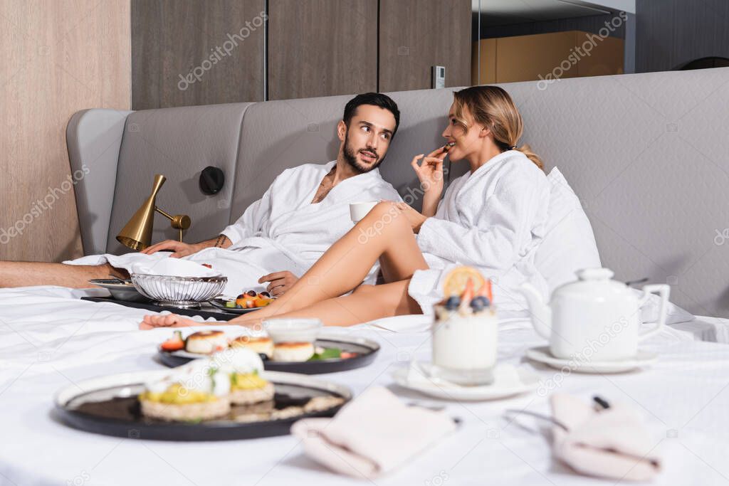 Young interracial couple in bathrobes sitting near tasty food on blurred foreground on hotel bed 