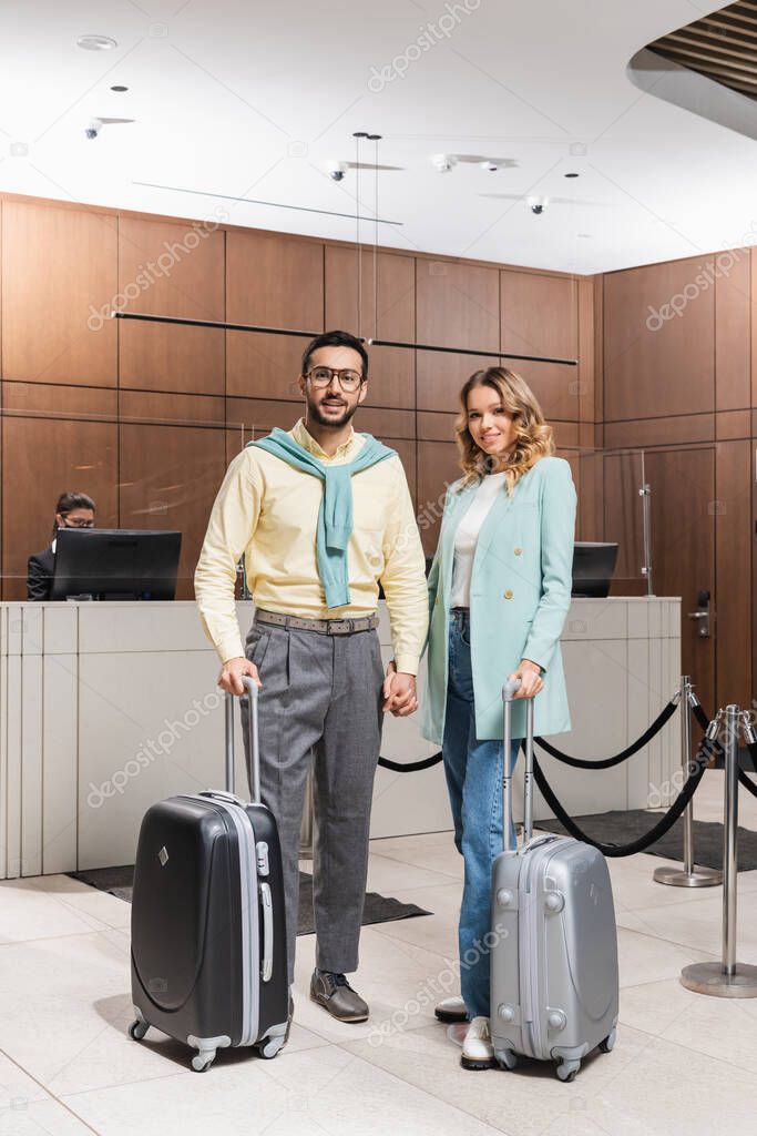 Smiling interracial couple with suitcases holding hands in hotel lobby 
