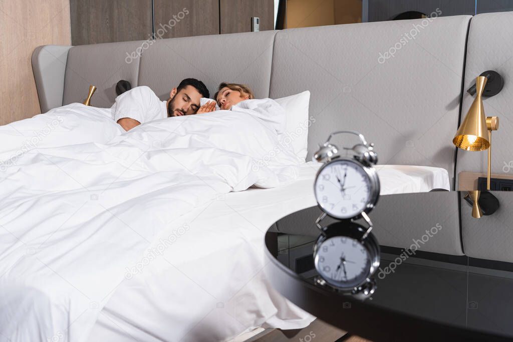 Interracial couple sleeping on white bedding near alarm clock on table on blurred foreground in hotel 