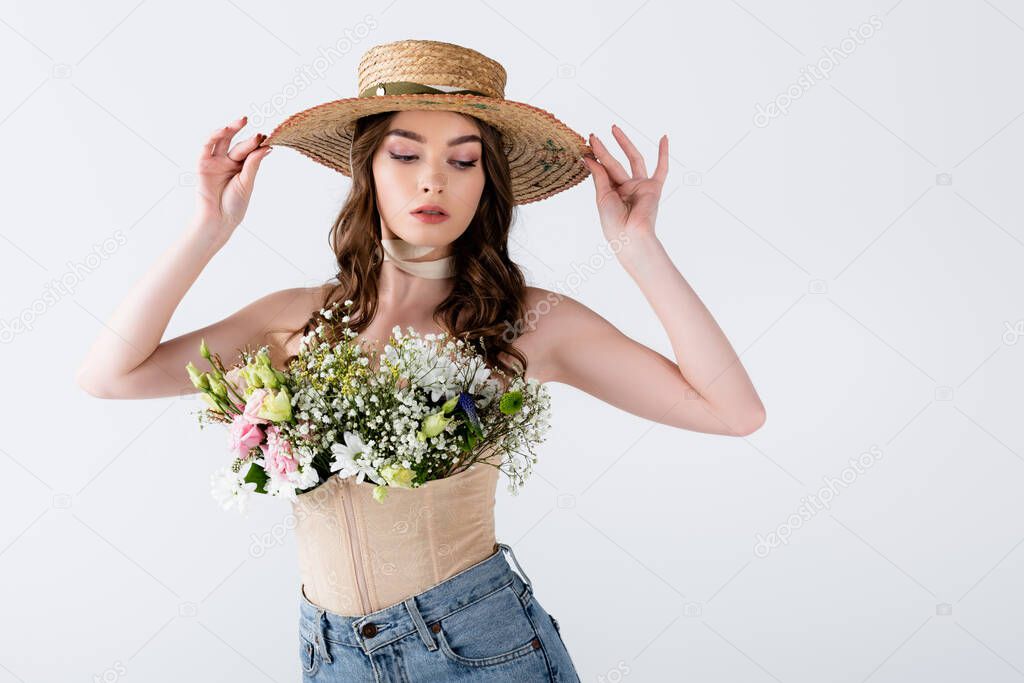 Trendy woman holding sun hat and posing with different flowers in blouse isolated on grey 