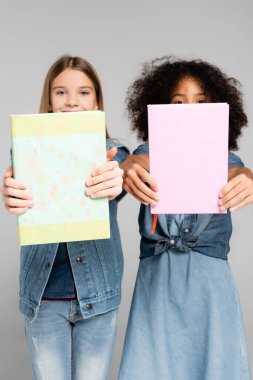 multicultural schoolgirls in denim clothes holding textbooks isolated on grey clipart
