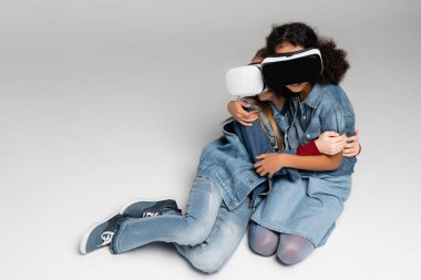 VR kulaklıklı korkmuş ırklar arası kızlar griye otururken kucaklaşıyorlar.