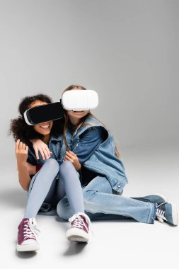 VR kulaklık takmış korkmuş ve heyecanlı çok kültürlü çocuklar gri renkte otururken kucaklaşıyorlar.