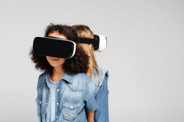 Gri renkte sırt sırta duran kot ve vr kulaklıklı moda ırklar arası kızlar.