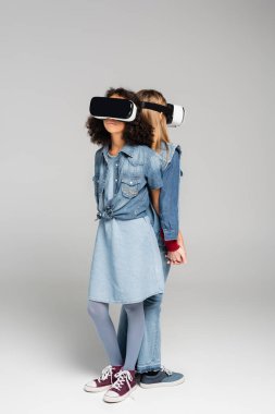 Gri renkte arka arkaya duran VR kulaklıklı modaya uygun ırklar arası arkadaşların tam boy görünümü