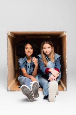 trendy interracial girls smiling at camera while sitting inside box on grey clipart
