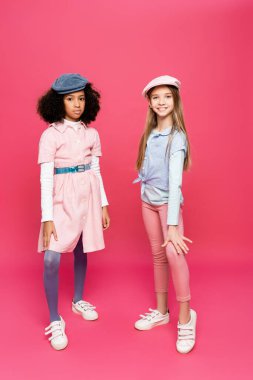 Moda giysiler, spor ayakkabılar ve şapkalar giymiş çocuklar pembe poz verirken kameraya bakıyorlar.